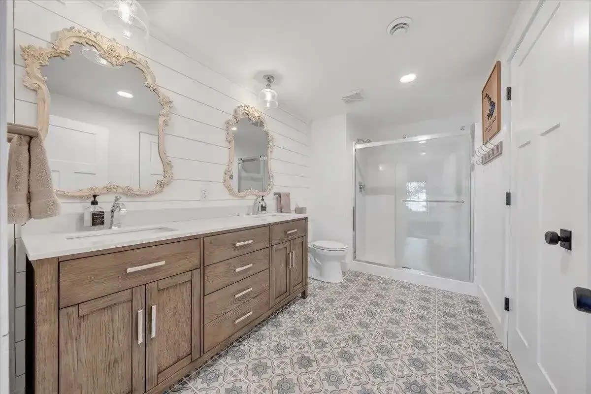 Bright bathroom with wood vanity, ornate mirror, patterned tile floor, and glass shower enclosure