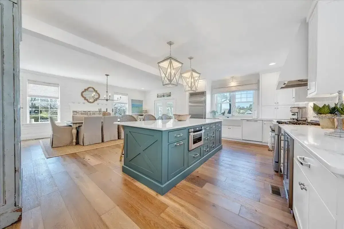 Bright white kitchen with teal island, hardwood floors, and dining area in the background