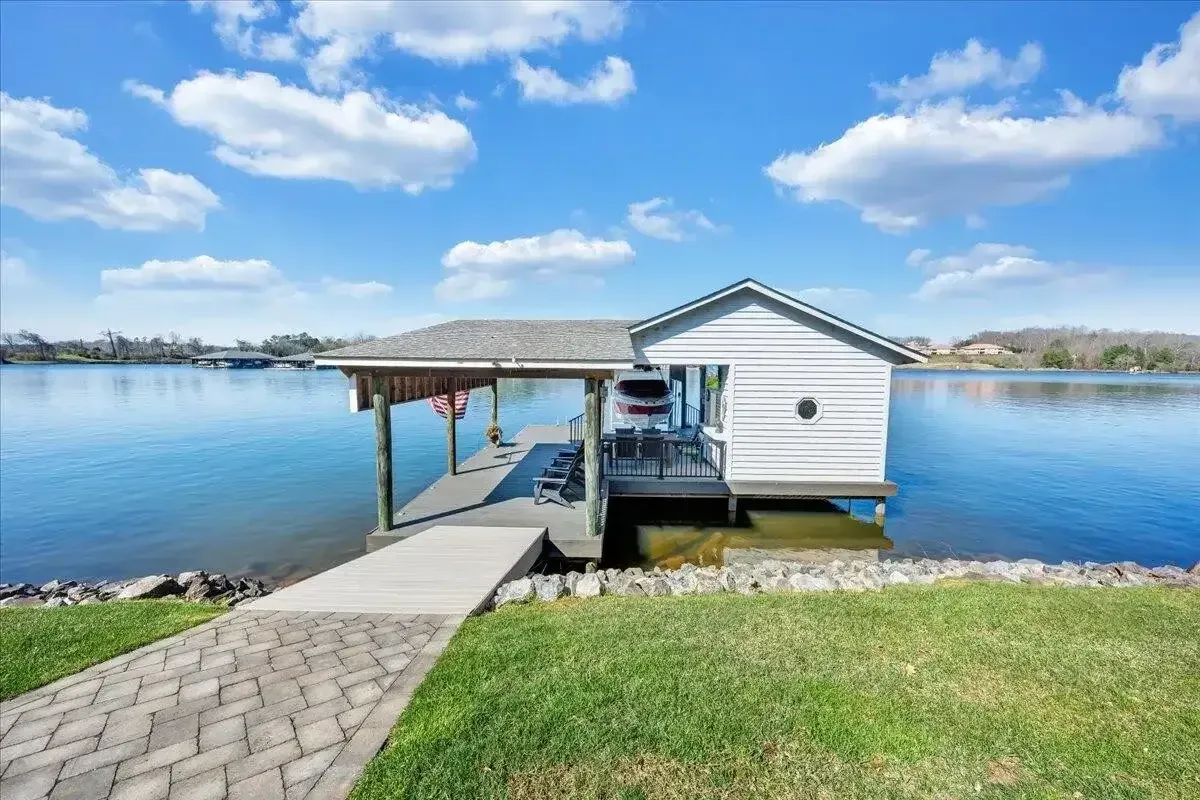 Small lakeside dock with a white boathouse, paved walkway, and calm blue water under a sunny sky