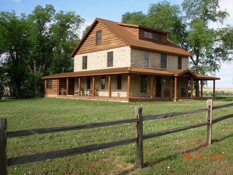 Rustic two-story log house with wraparound porch and a fenced lawn, surrounded by trees under a clear sky.