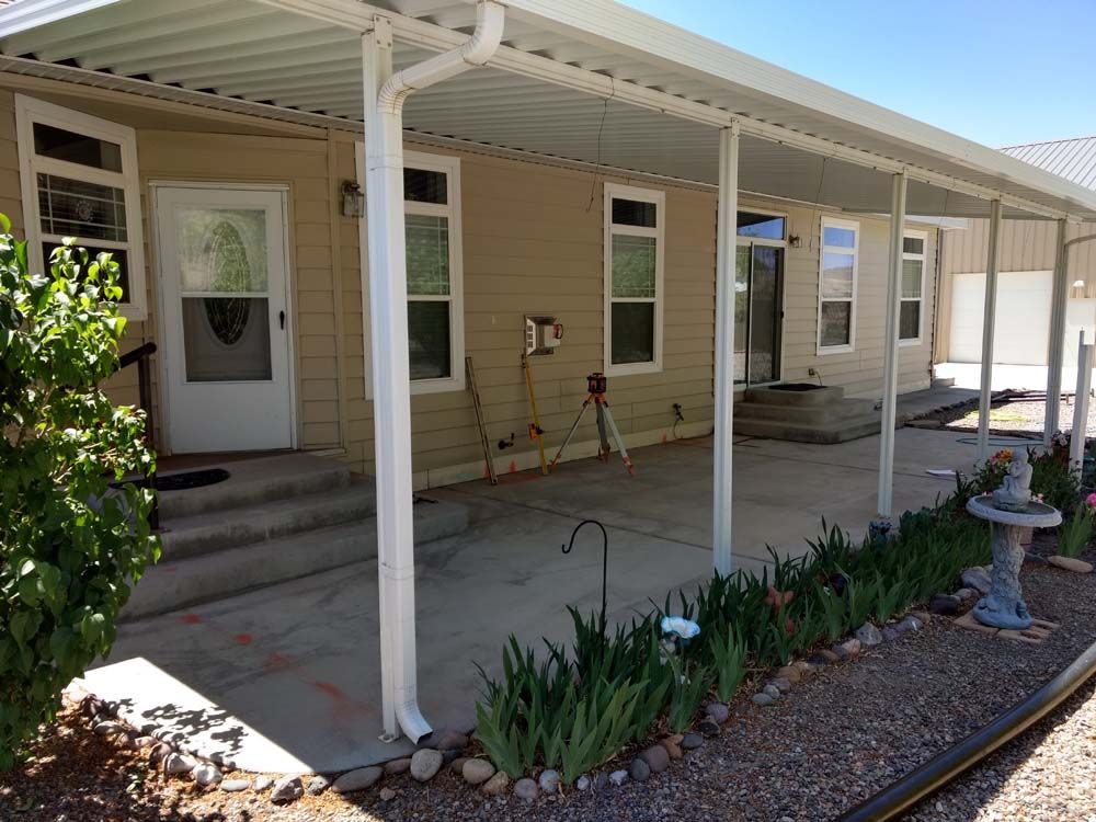 Covered front porch of a beige house with white columns, potted plants, and a walkway garden bed.