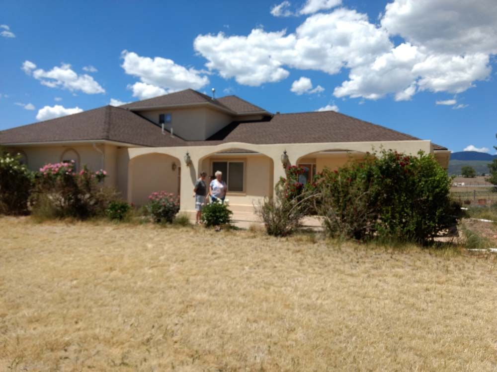 Single-story house with arched porch, dry front yard, and two people standing by the entrance under a blue sky.