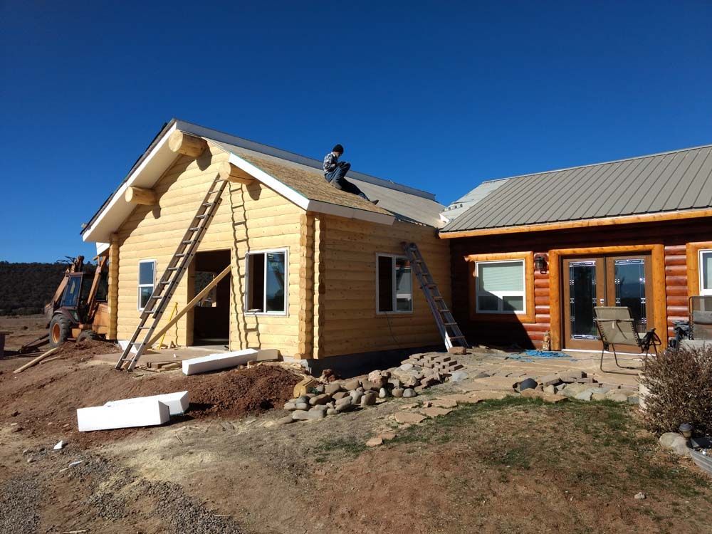 Two workers on ladders building a small tan house beside a completed wooden home under a clear blue sky