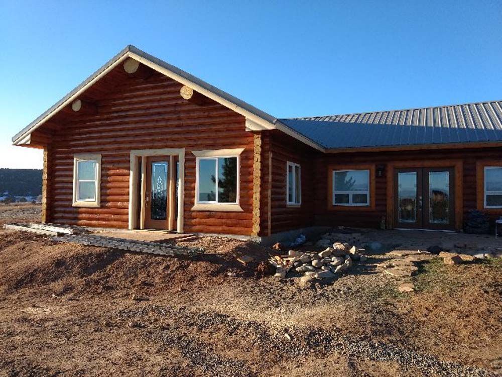 Log cabin-style house with log siding and a metal roof on a bare dirt lot under a blue sky