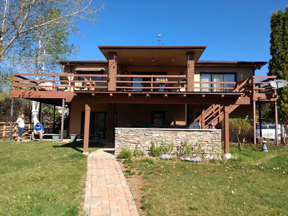 Two-story brown house with a wooden deck and stone planter, viewed from a front walkway under a blue sky