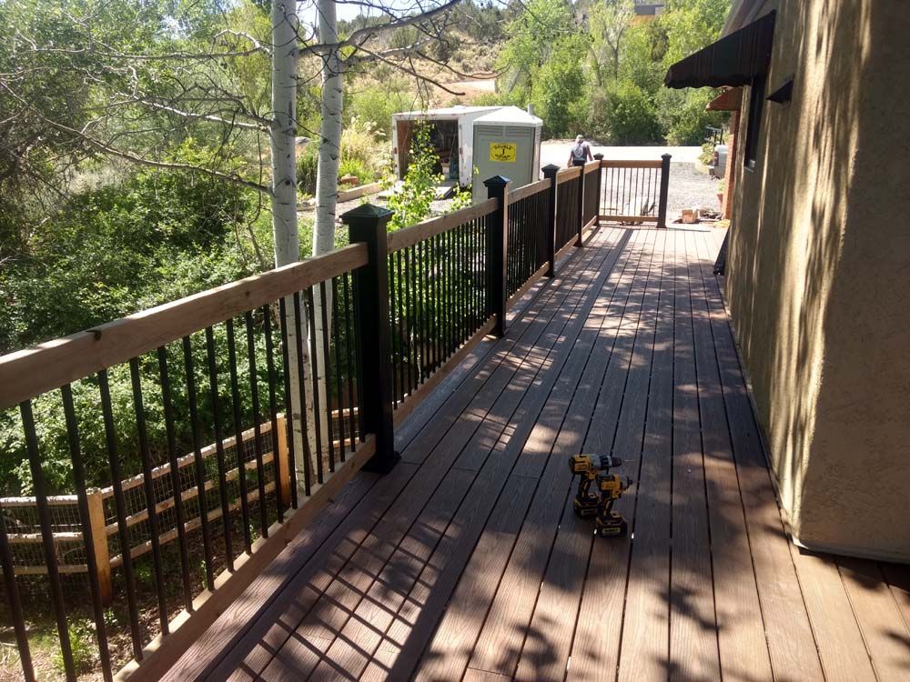 Wooden deck walkway beside a tan building, with black railing and trees below in bright sunlight.