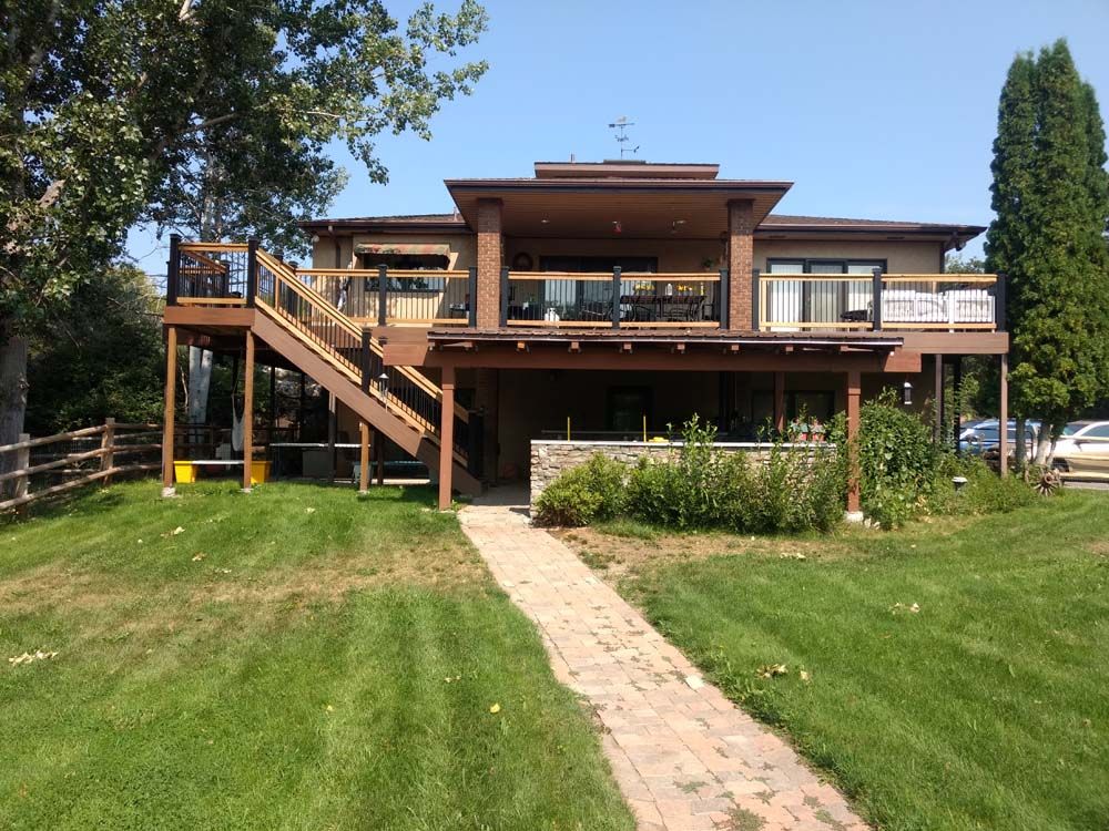Wooden house with wraparound deck and stairs, viewed from a grassy yard with a stone walkway