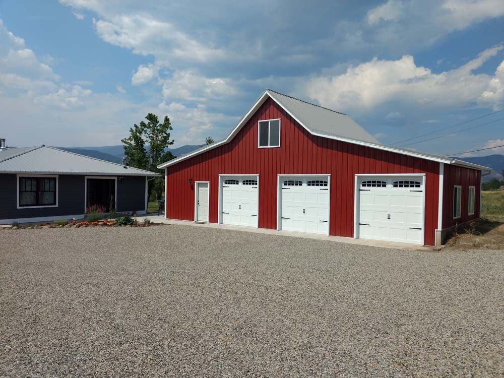 Red barn with three white garage doors beside a small house under a cloudy sky.