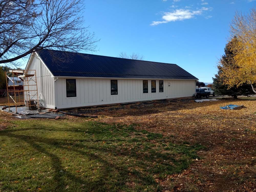 White house with dark roof in a grassy yard under a blue sky, with autumn trees nearby