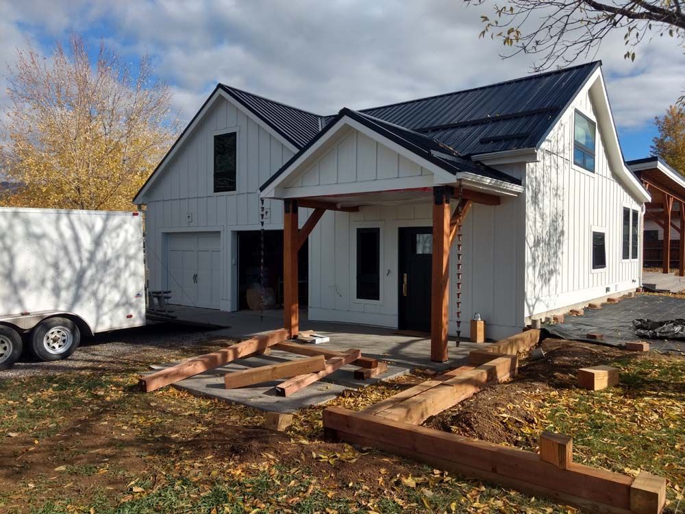 Partially built white house with black roof and front porch in a yard with lumber and trailer
