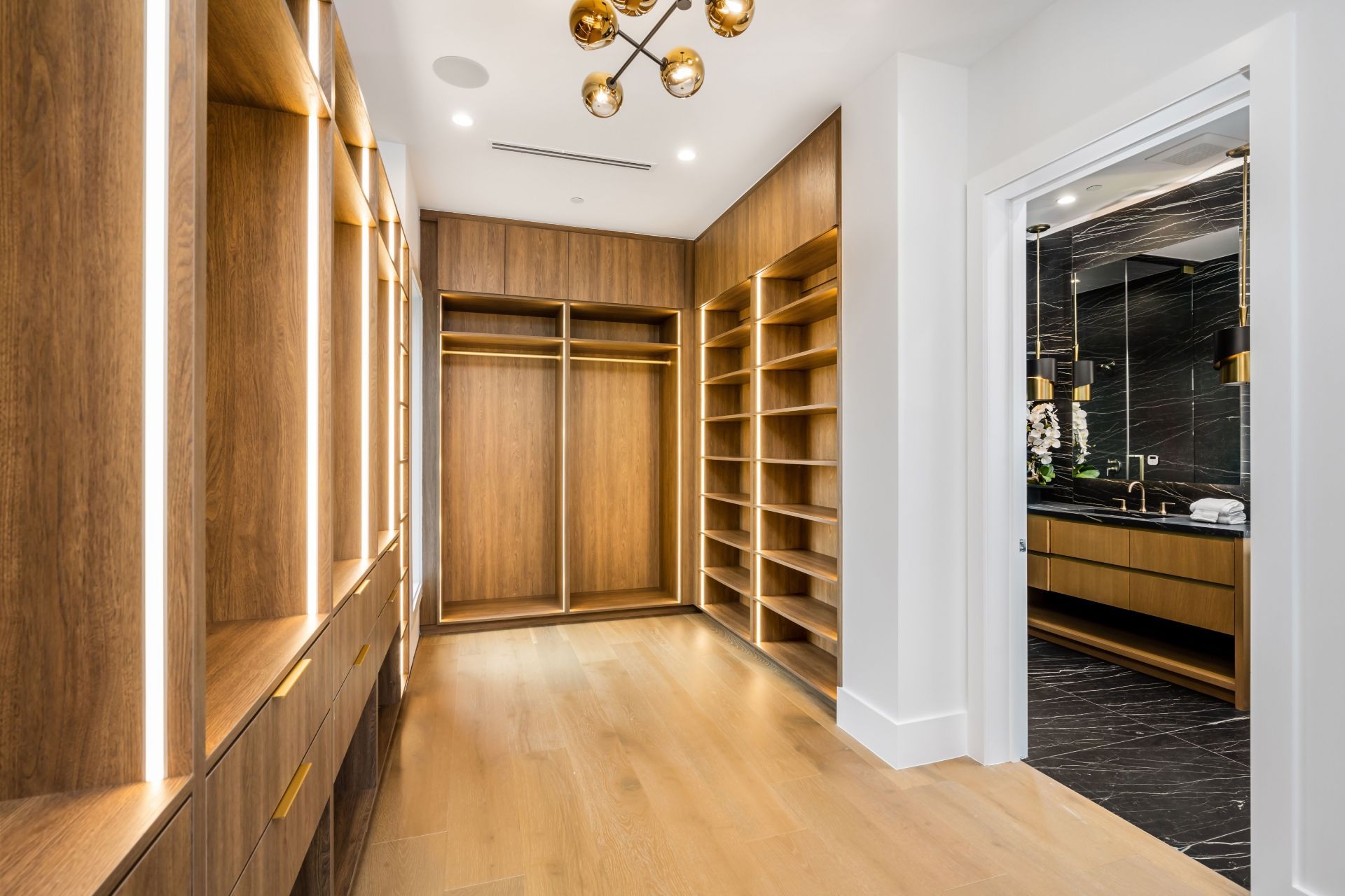 Spacious walk-in closet with light wood shelves, hanging space, and an open doorway to a dark-tiled bathroom.