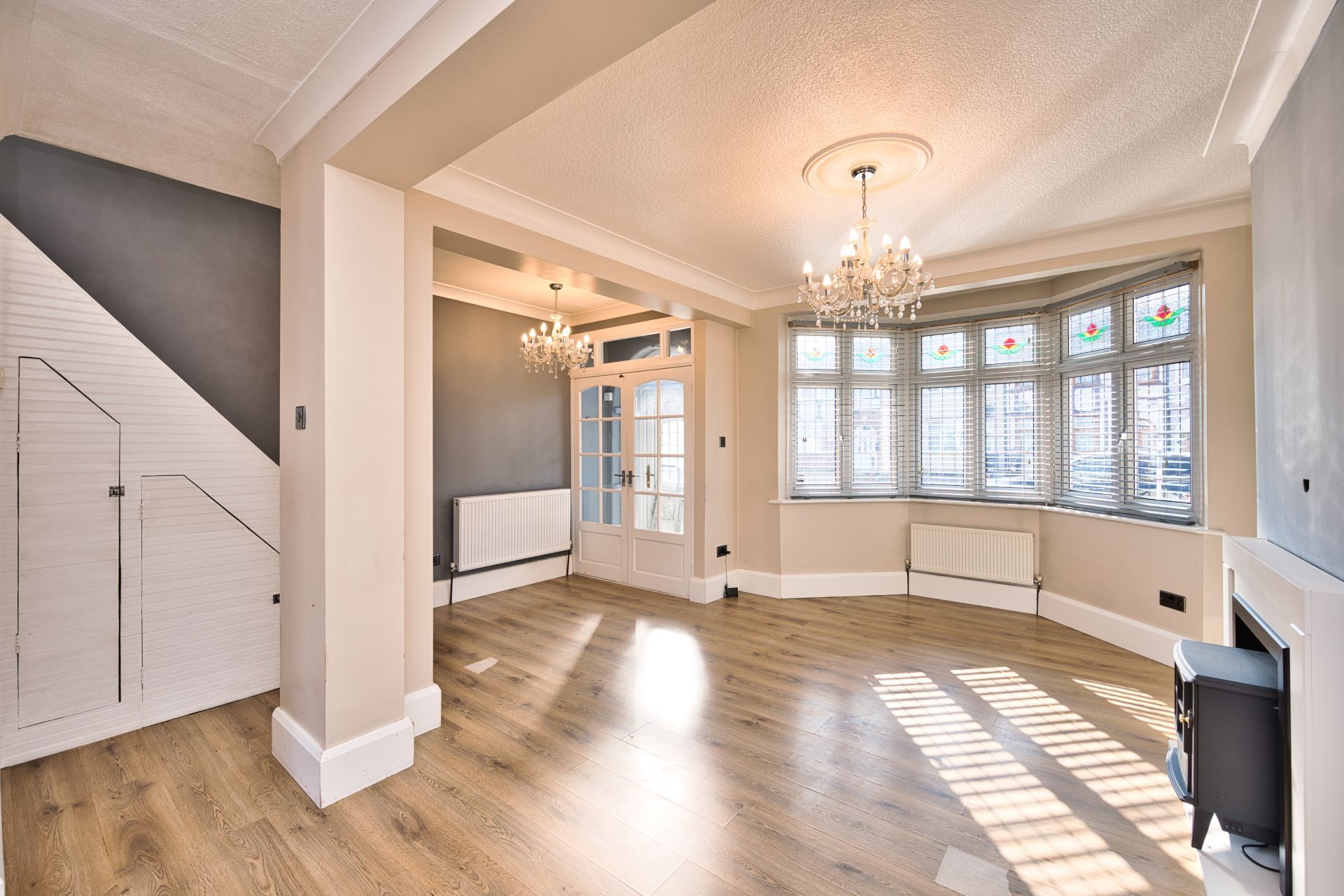 Bright empty living room with bay windows, chandelier, and light wood floors, sunlight streaming in