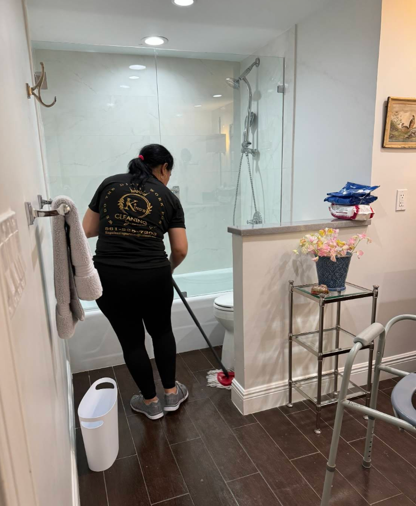 Person cleaning a bathroom floor with a mop. White and gray bathroom with glass shower.