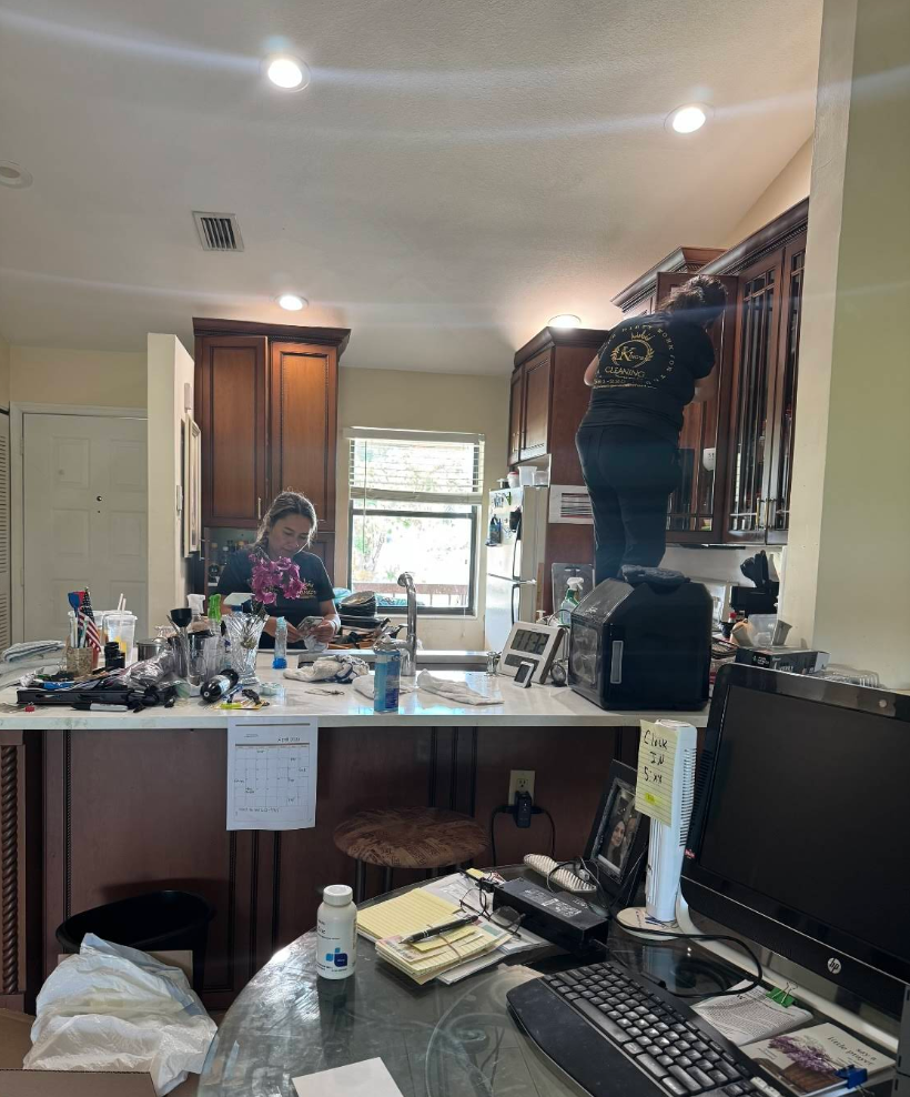 Two people working in a kitchen; one on a ladder reaching up, the other at a counter with items.