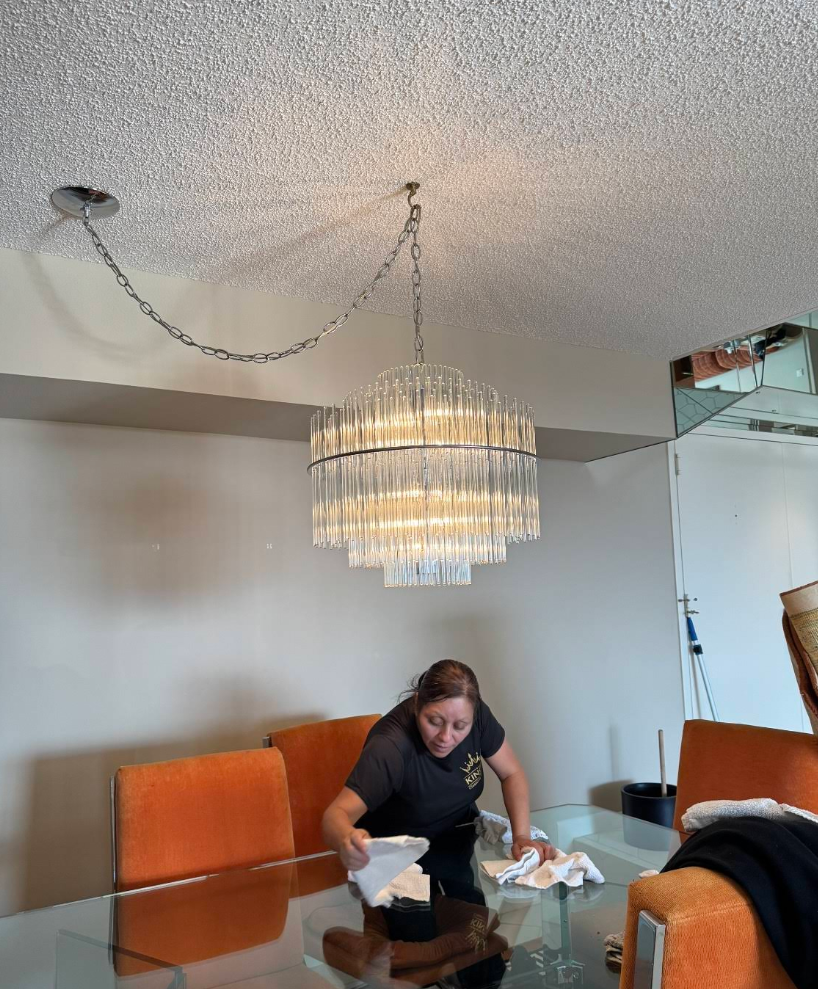 Woman cleaning a glass dining table under a large chandelier with orange chairs.