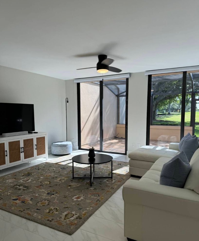 Living room with a beige sectional, TV, and black coffee table. Sliding glass doors lead to a patio with outdoor space.