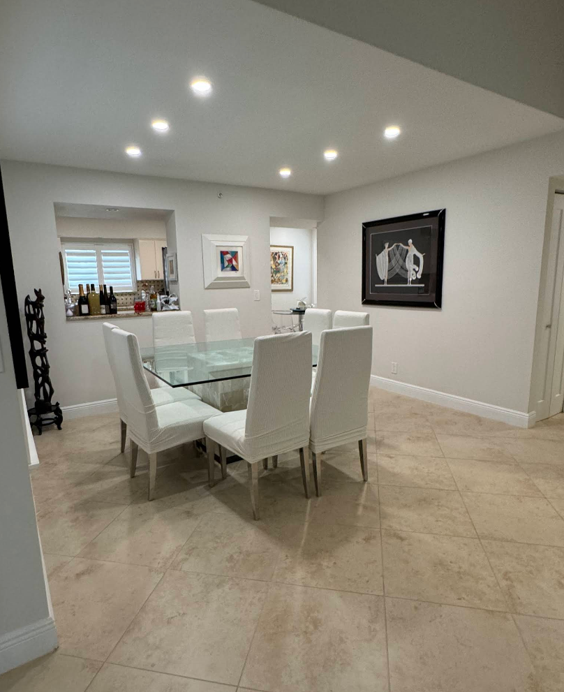 Dining room with glass table, white chairs, and artwork on light walls. Beige tiled floor.
