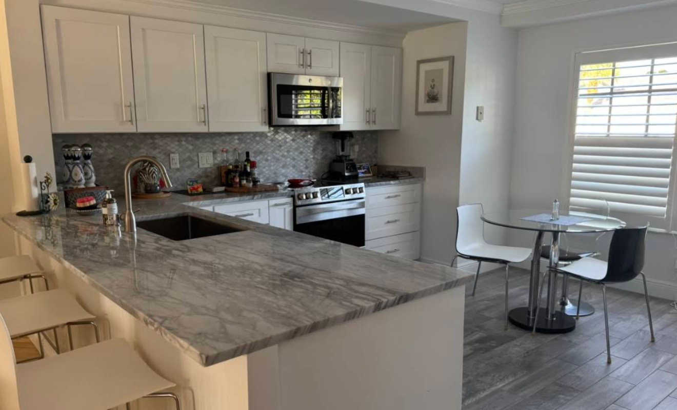 White kitchen with a marble countertop island and stainless steel appliances. A small table and chairs sit by the window.