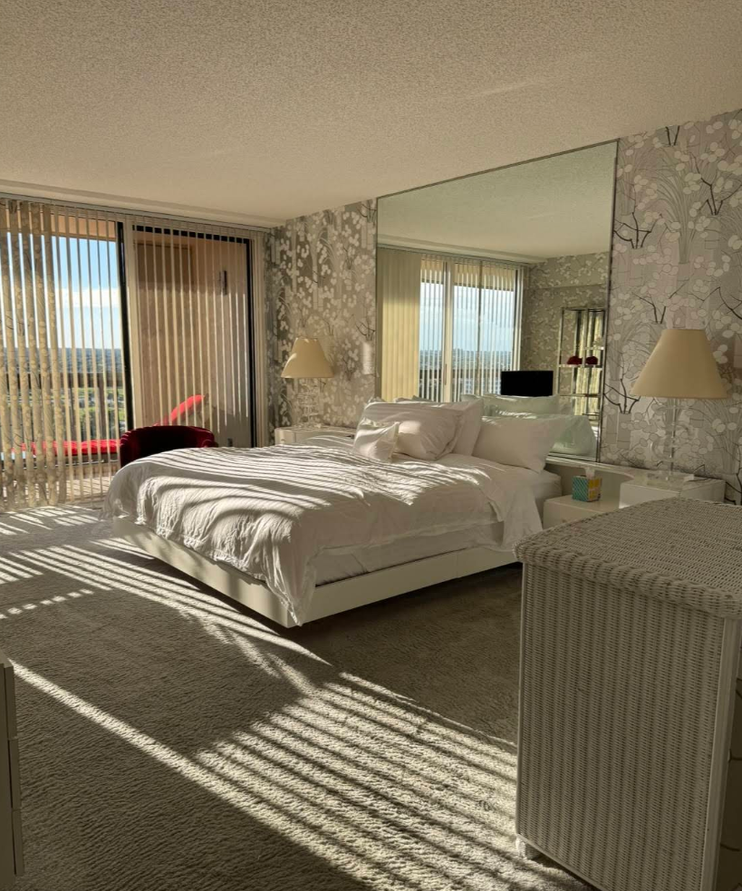 Bedroom with a white bed, floral wallpaper, large mirror, and sunlight streaming through blinds.