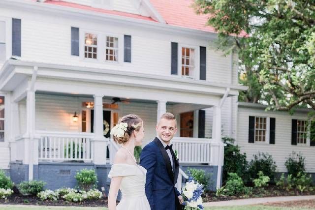 Bride and groom walking in front of a white house with a porch; he is smiling, and she is looking back.