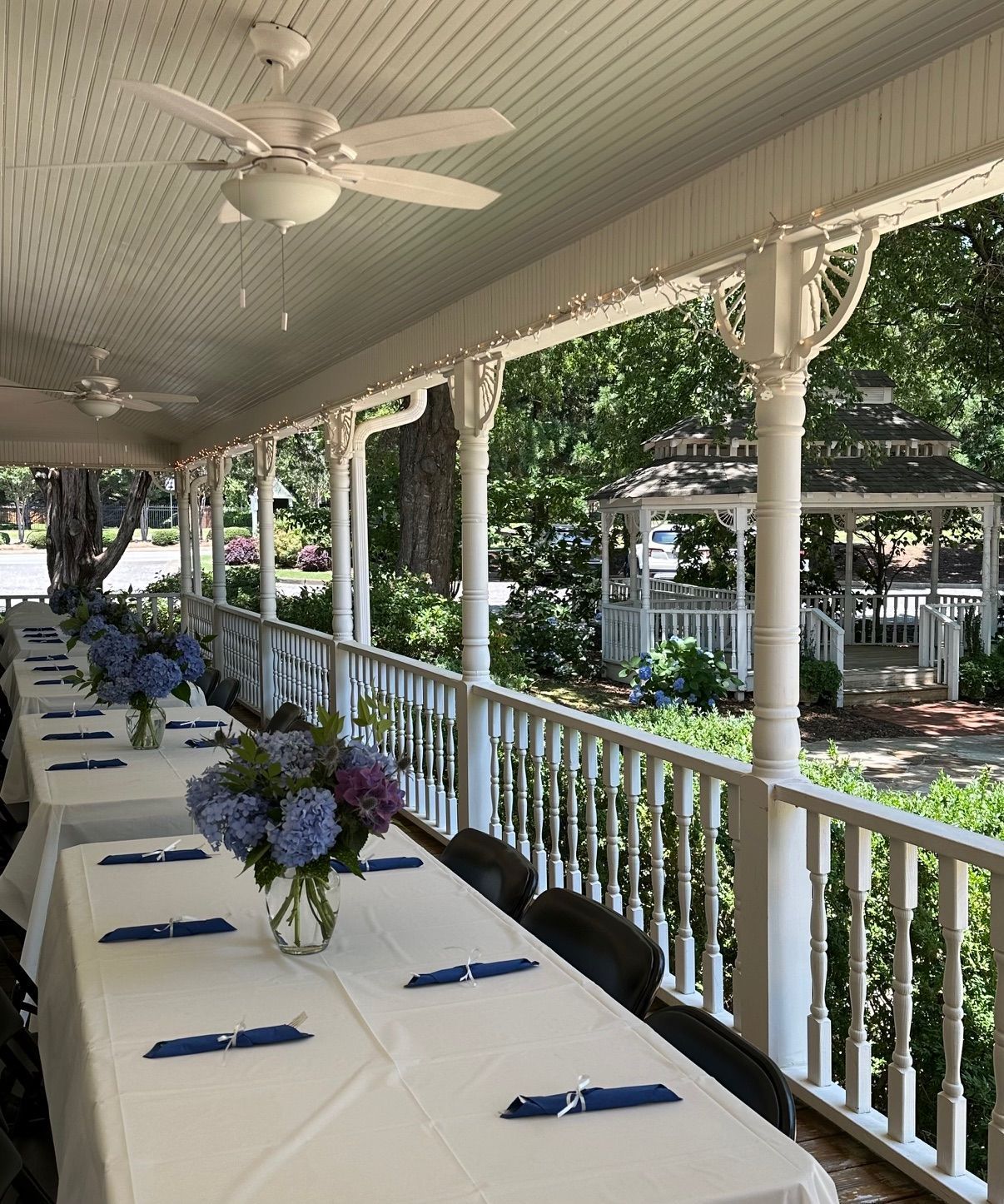 Long tables set for a gathering on a porch, blue flowers, gazebo in the background.