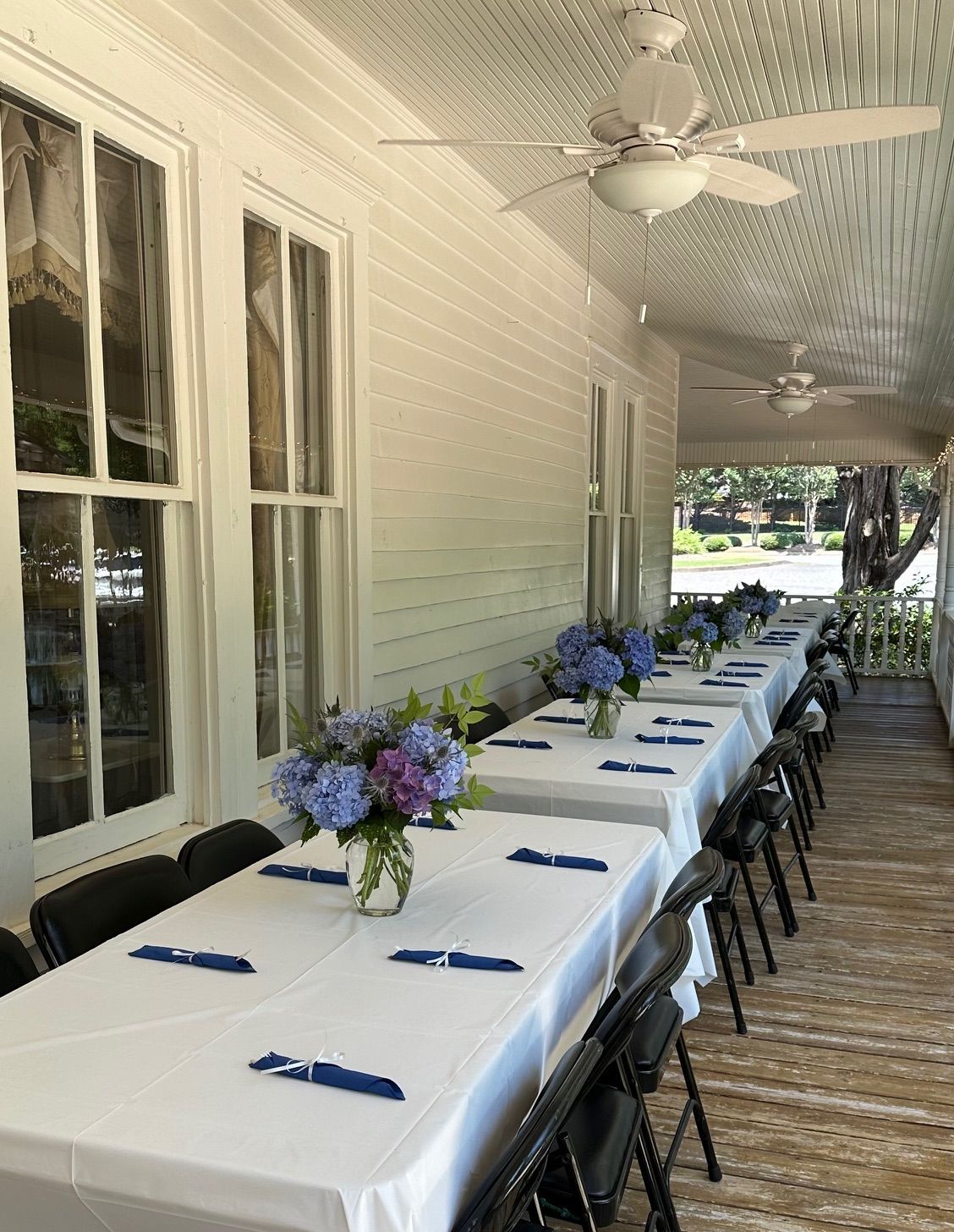 Long tables set with white tablecloths and blue accents, decorated with flowers on a white porch.