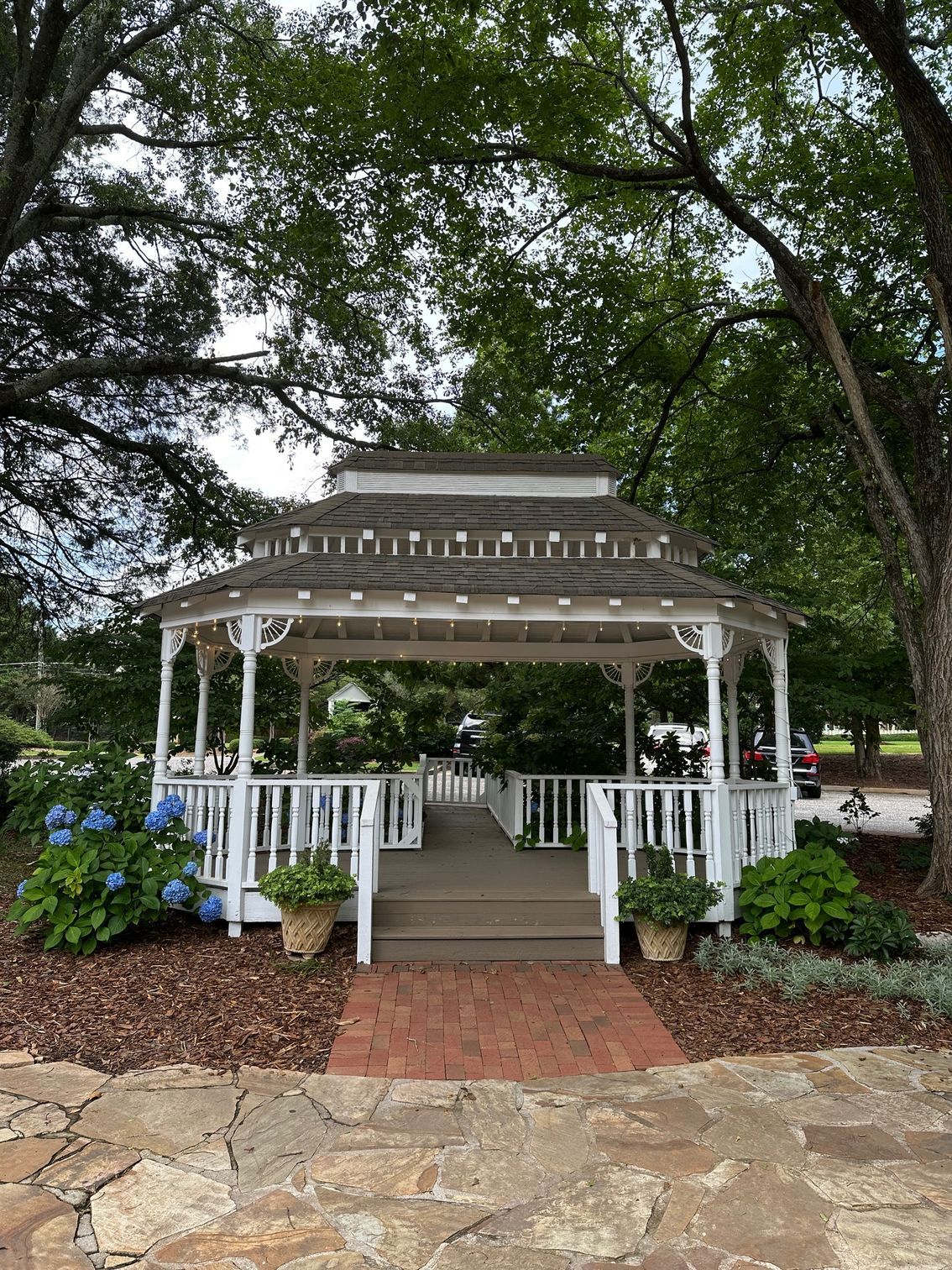 White gazebo with steps, surrounded by plants and trees, located in a park setting.