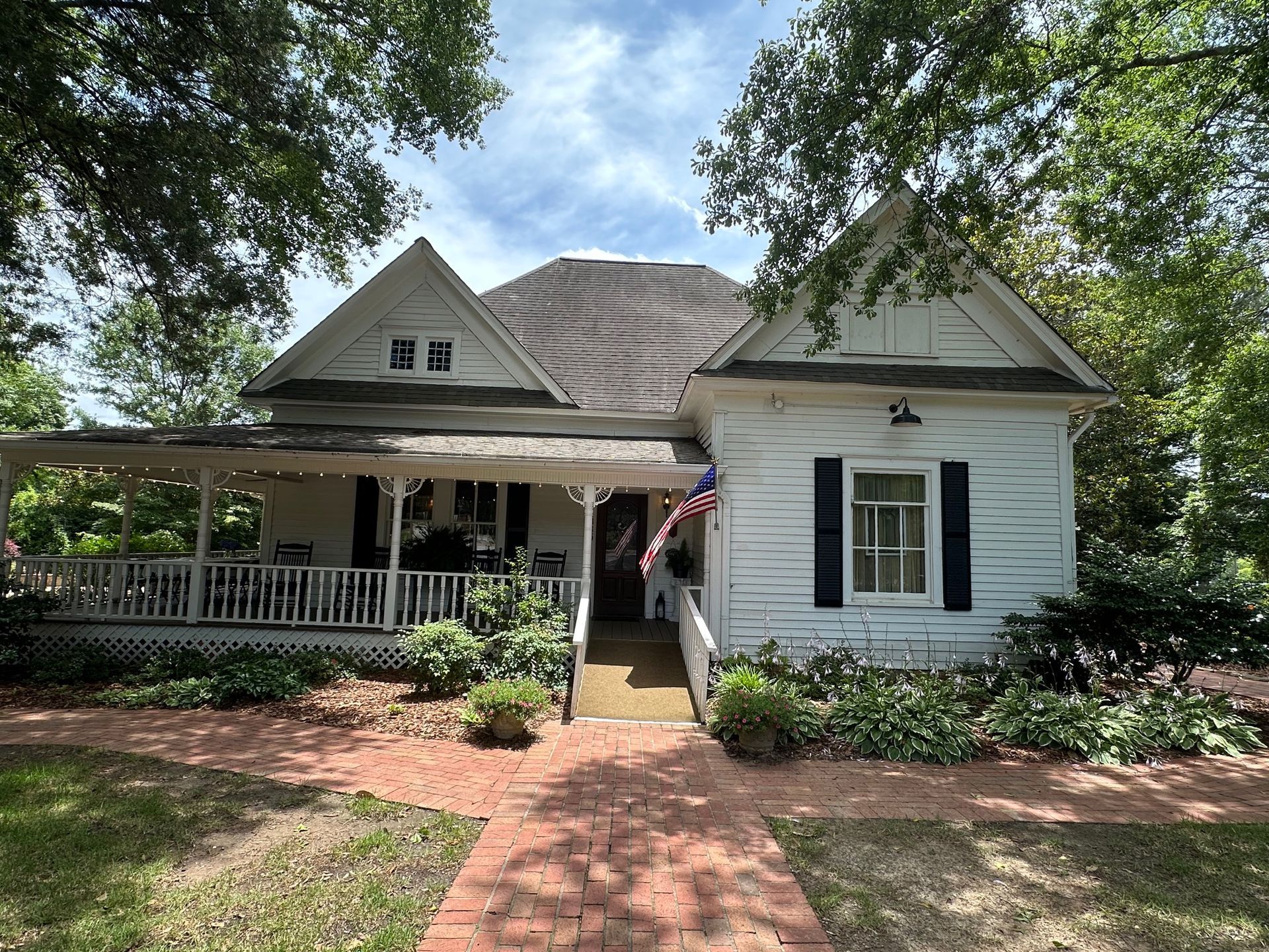 White cottage with a wraparound porch and brick walkway, surrounded by greenery under a partly cloudy sky.