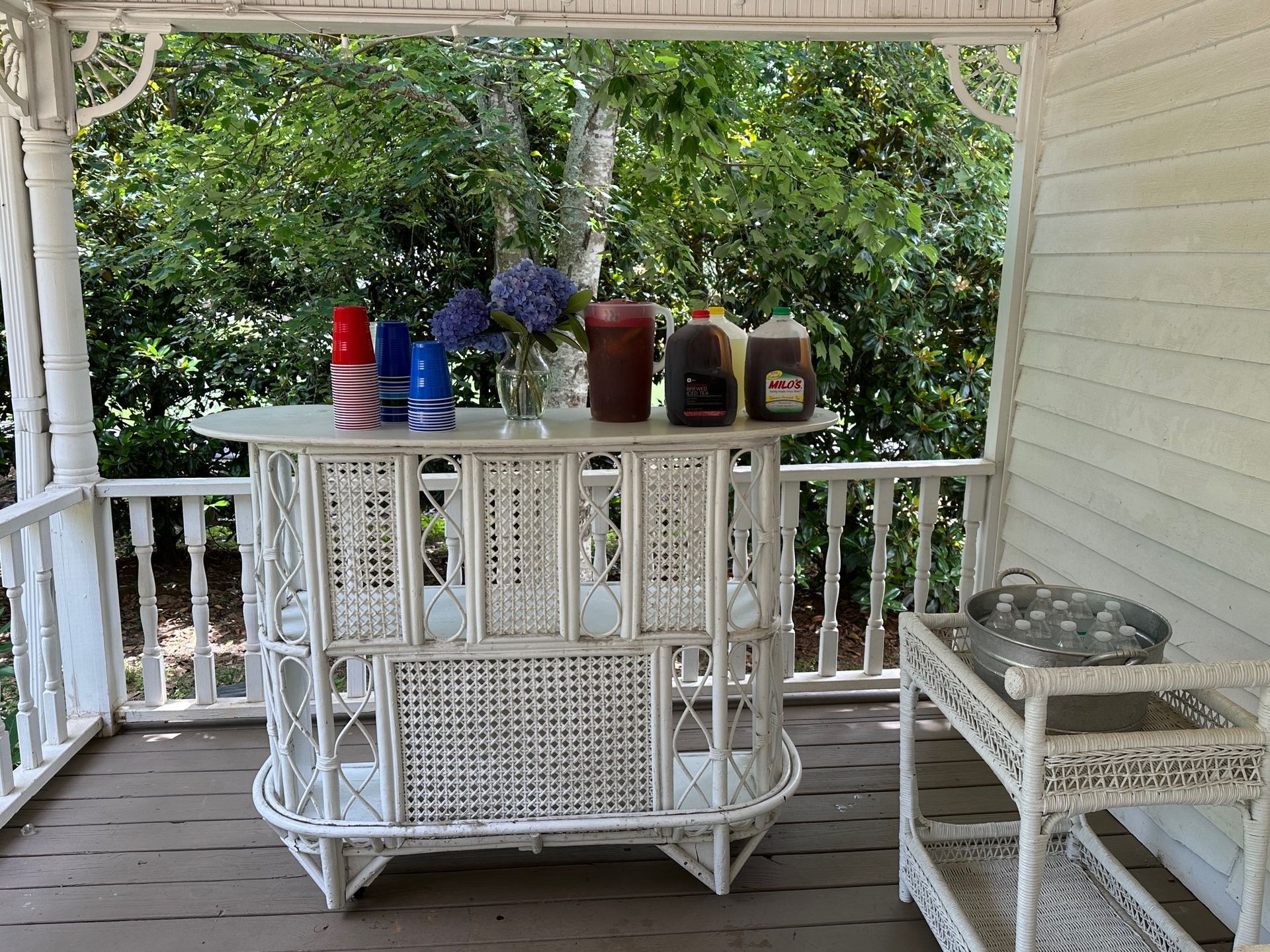 White wicker bar on a porch, drinks and flowers on top, surrounded by railings, with trees in the background.