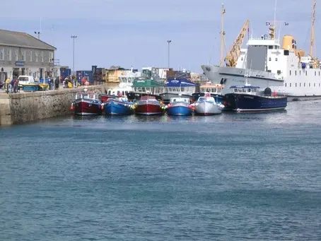 Boats docked in a harbor, next to a stone wall and buildings, with a large white ship in the background.