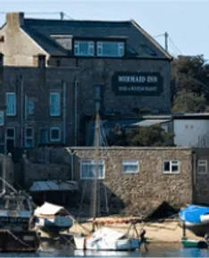 Mermaid Inn pub by a harbor. Stone buildings, a sailboat, and boats in the water on a sunny day.