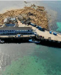Harbor with buildings, a pier, boats, and a rocky island. Blue water surrounds it.