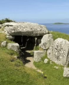 Prehistoric stone structure, capstone supported by upright stones, grassy hillside, ocean in background.