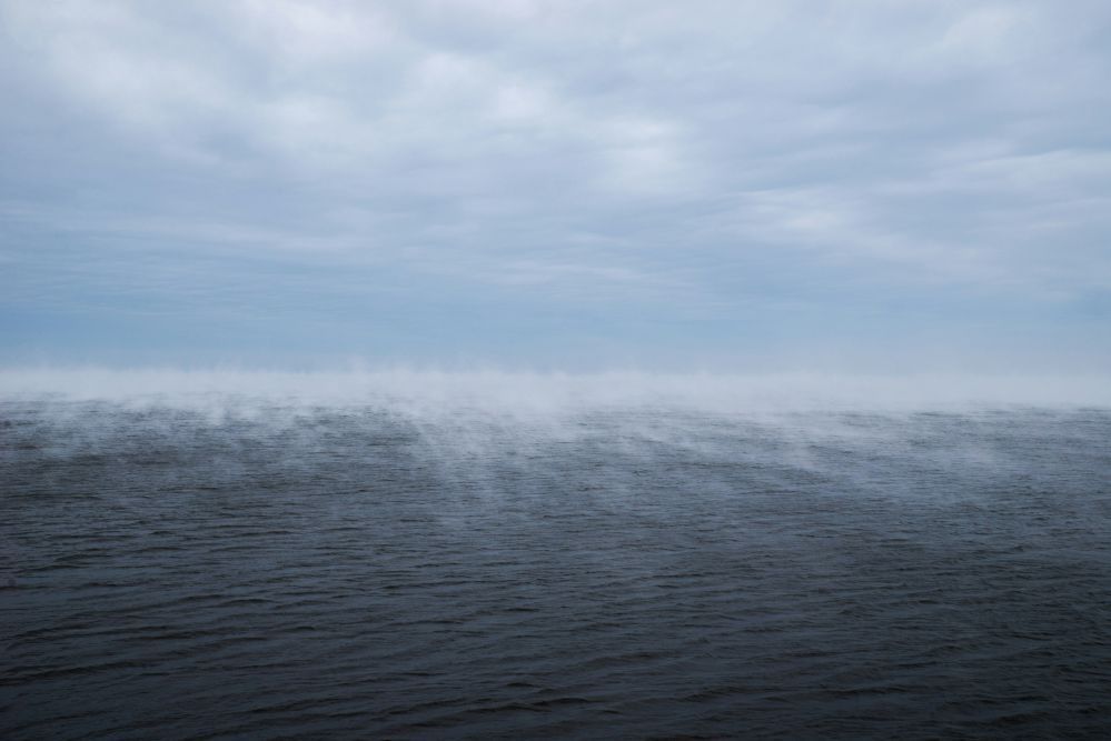 Dark water with white mist rising to a pale blue overcast sky.
