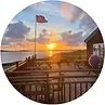 Sunset view from a deck; ocean, flagpole with flag, house, and cloudy sky.