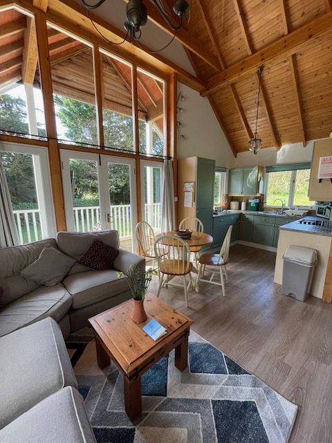 Open-plan cabin interior with large windows. A sofa, table, chairs, and kitchen are visible.