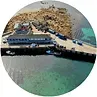 Aerial view of a pier with buildings, cars, and boats on blue-green water; rocky island background.