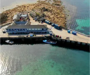 Coastal pier with building, boats, vehicles, and rocky island. Turquoise water surrounds.