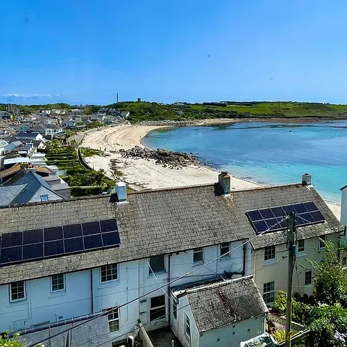 View of beach and town from rooftop, featuring solar panels on houses, sandy shore, and clear blue water.