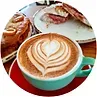 Latte with heart-shaped foam art, pastries, and a sandwich on a table.