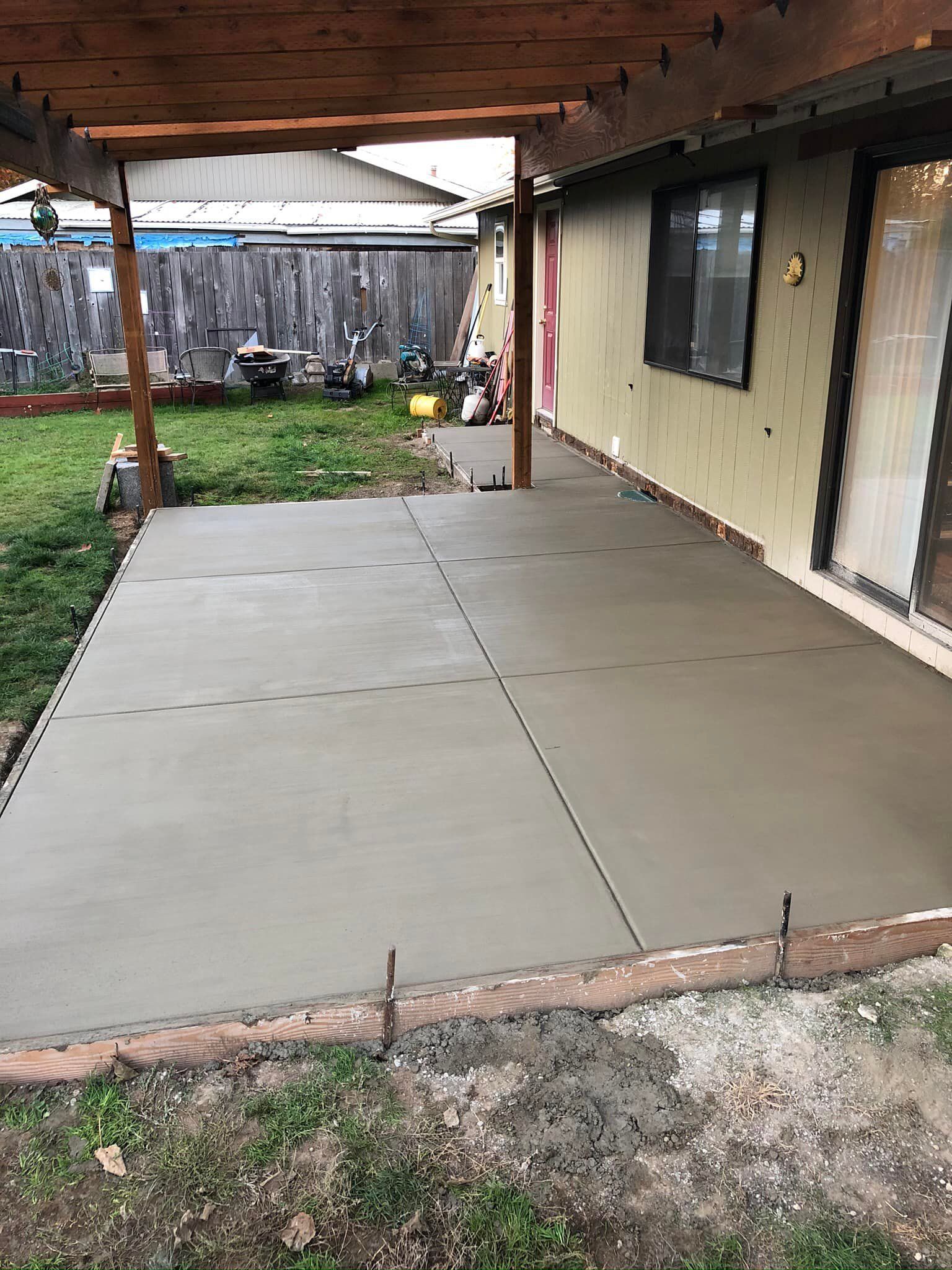 A newly poured concrete patio extending from a house wall under a wooden pergola, viewed from the backyard lawn.