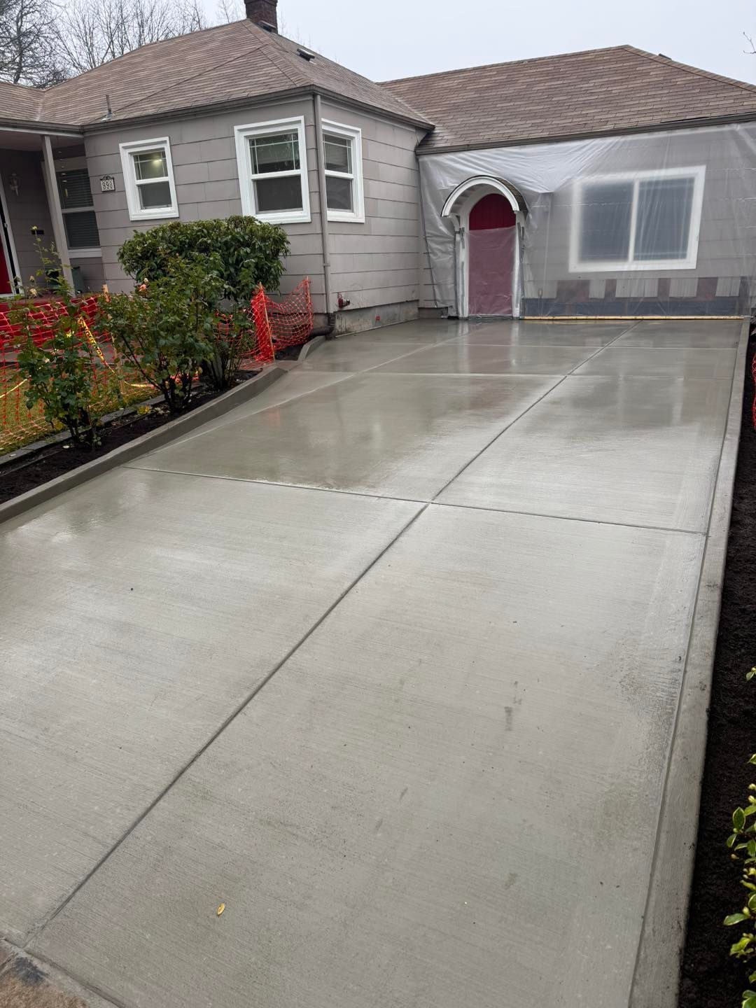 A newly poured, wet concrete driveway in front of a gray house with a prominent arched red front door.