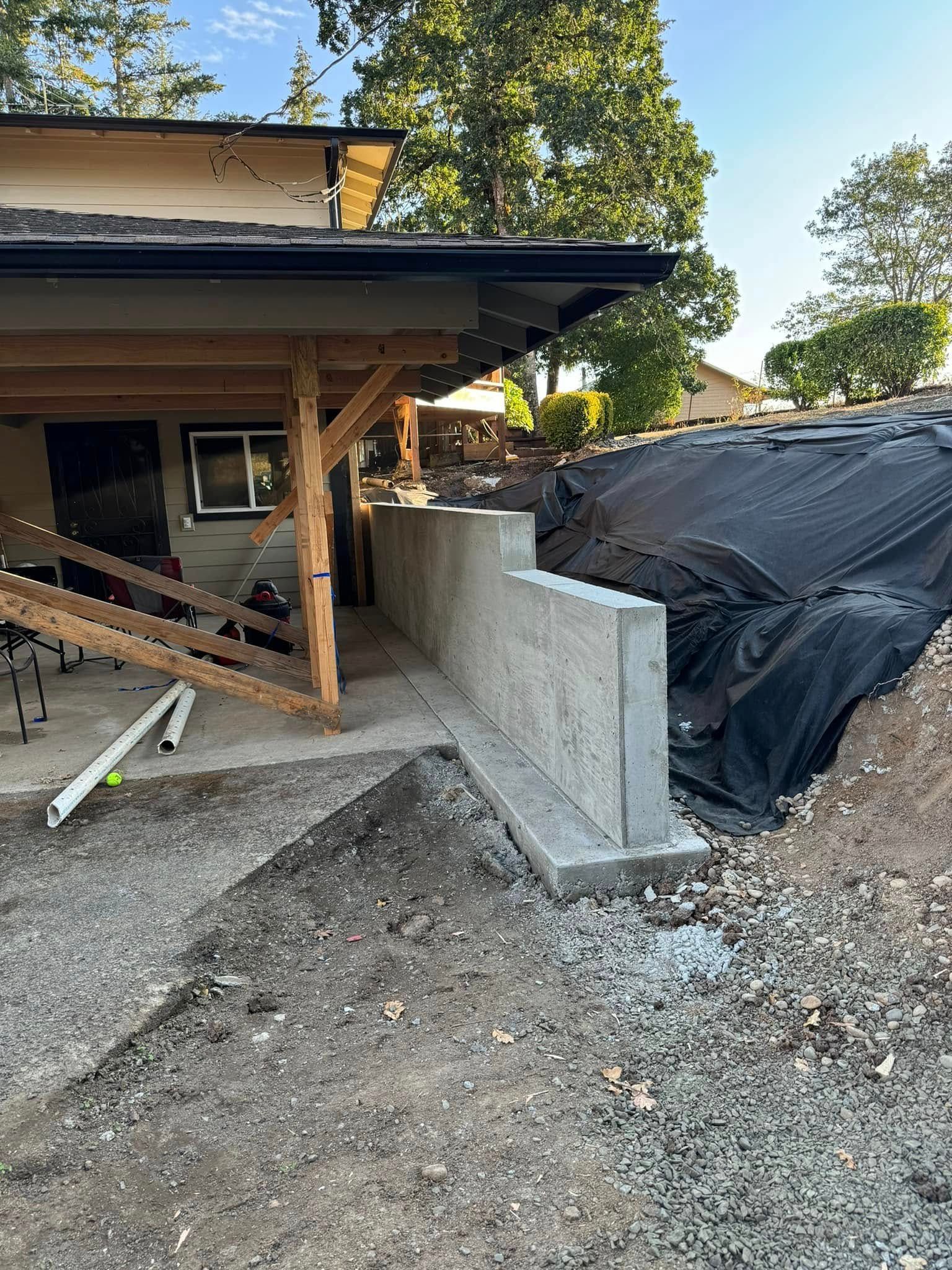 A newly poured concrete retaining wall sits on gravel next to a house with an attached wooden-framed covered porch area.