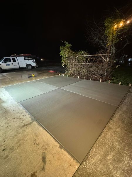 A freshly poured square concrete slab sits on a driveway at night, with a white service truck parked in the background.