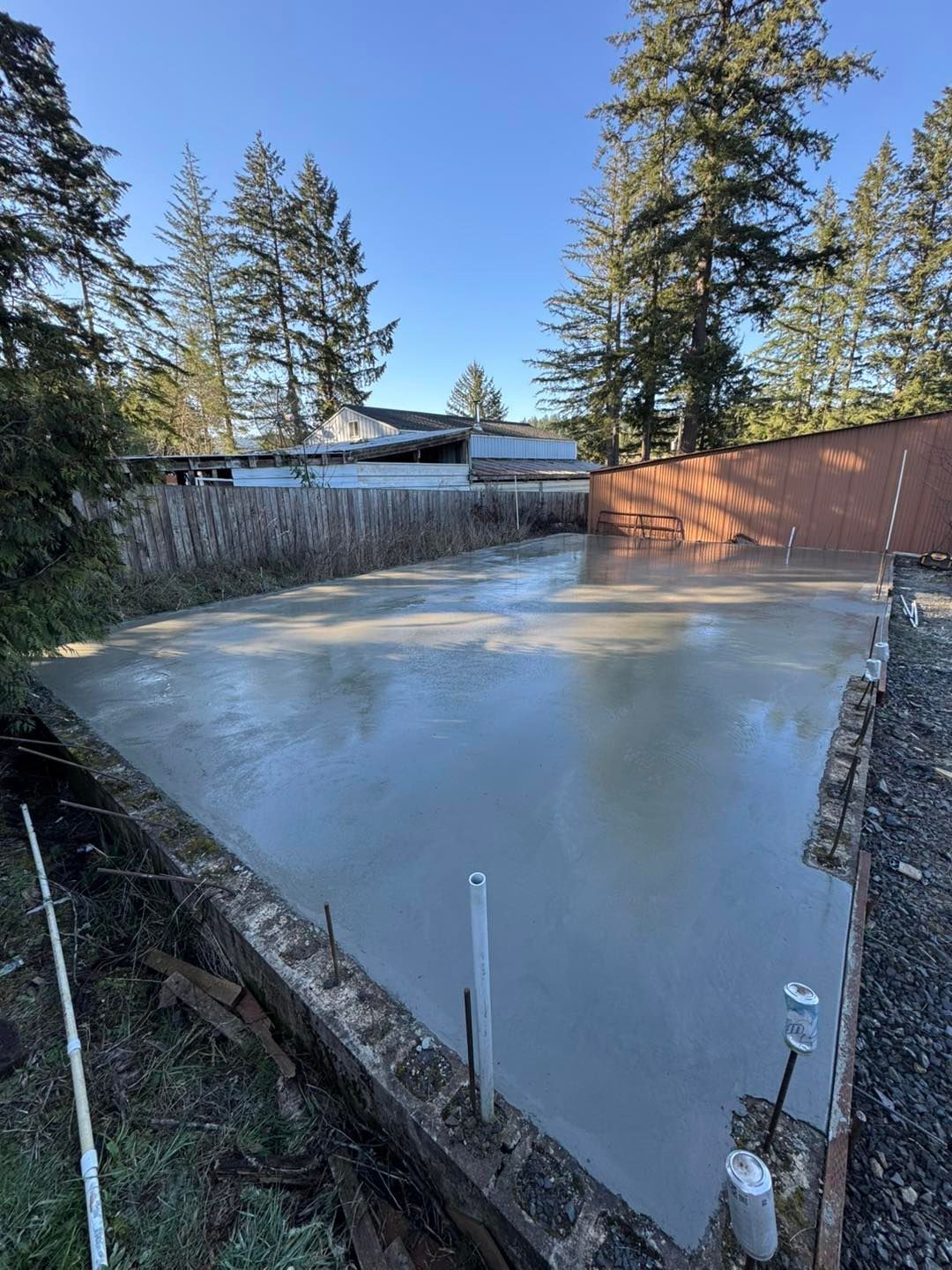 A freshly poured concrete slab foundation sits in a yard surrounded by wooden fences and tall trees.