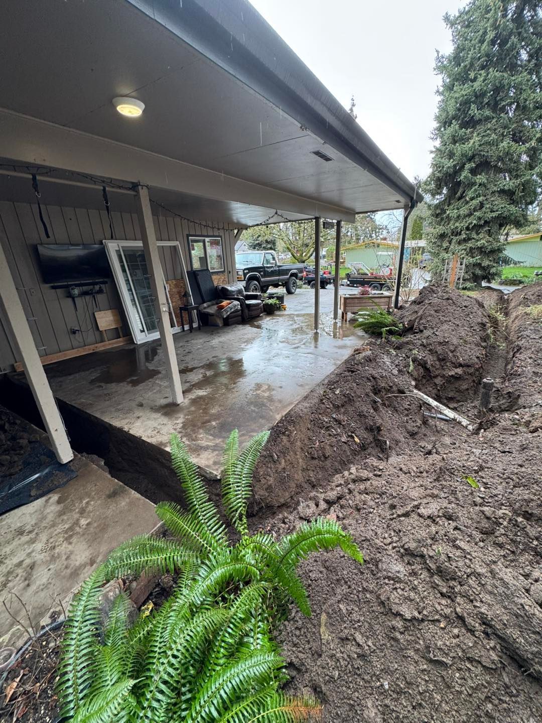 A covered patio with construction trenching in the yard and a patio door leaning against the exterior wall.