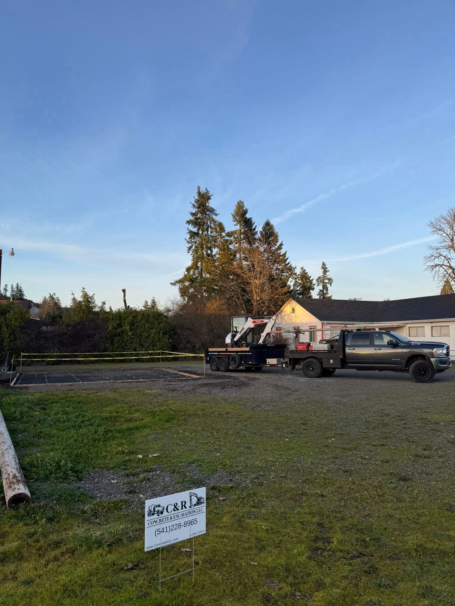 A truck pulls a trailer with a piece of heavy equipment on a gravel lot next to a grassy area with a small sign.