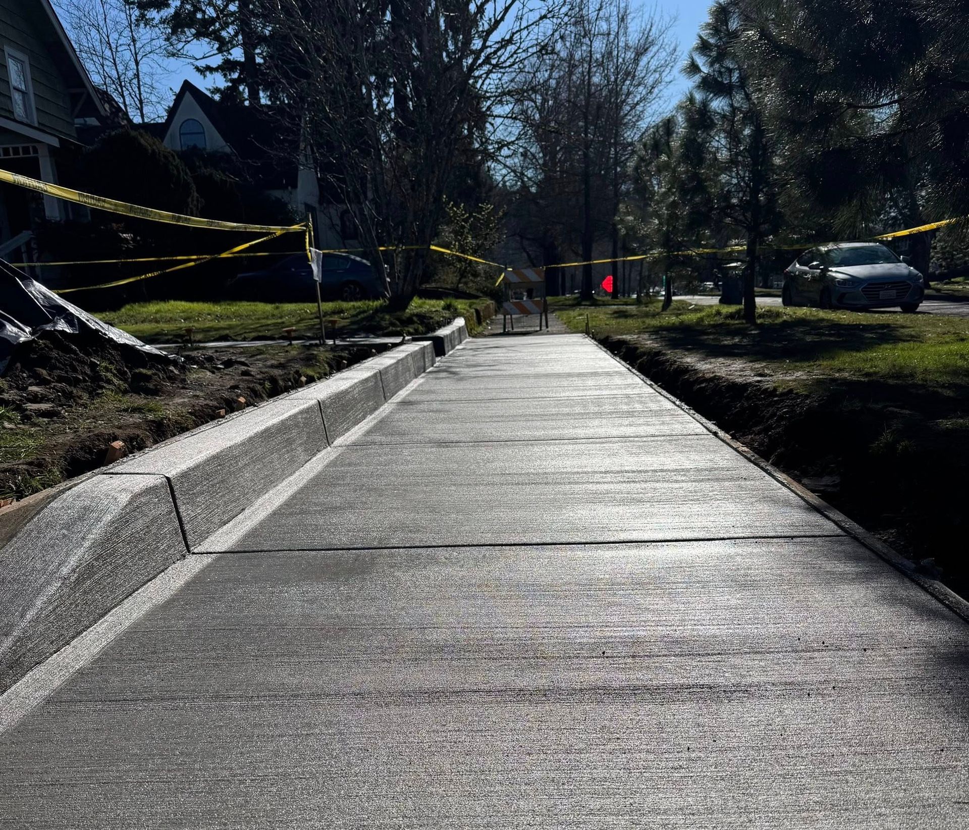 A newly poured grey concrete sidewalk with a slanted curb, bordered by yellow caution tape on a sunny street.