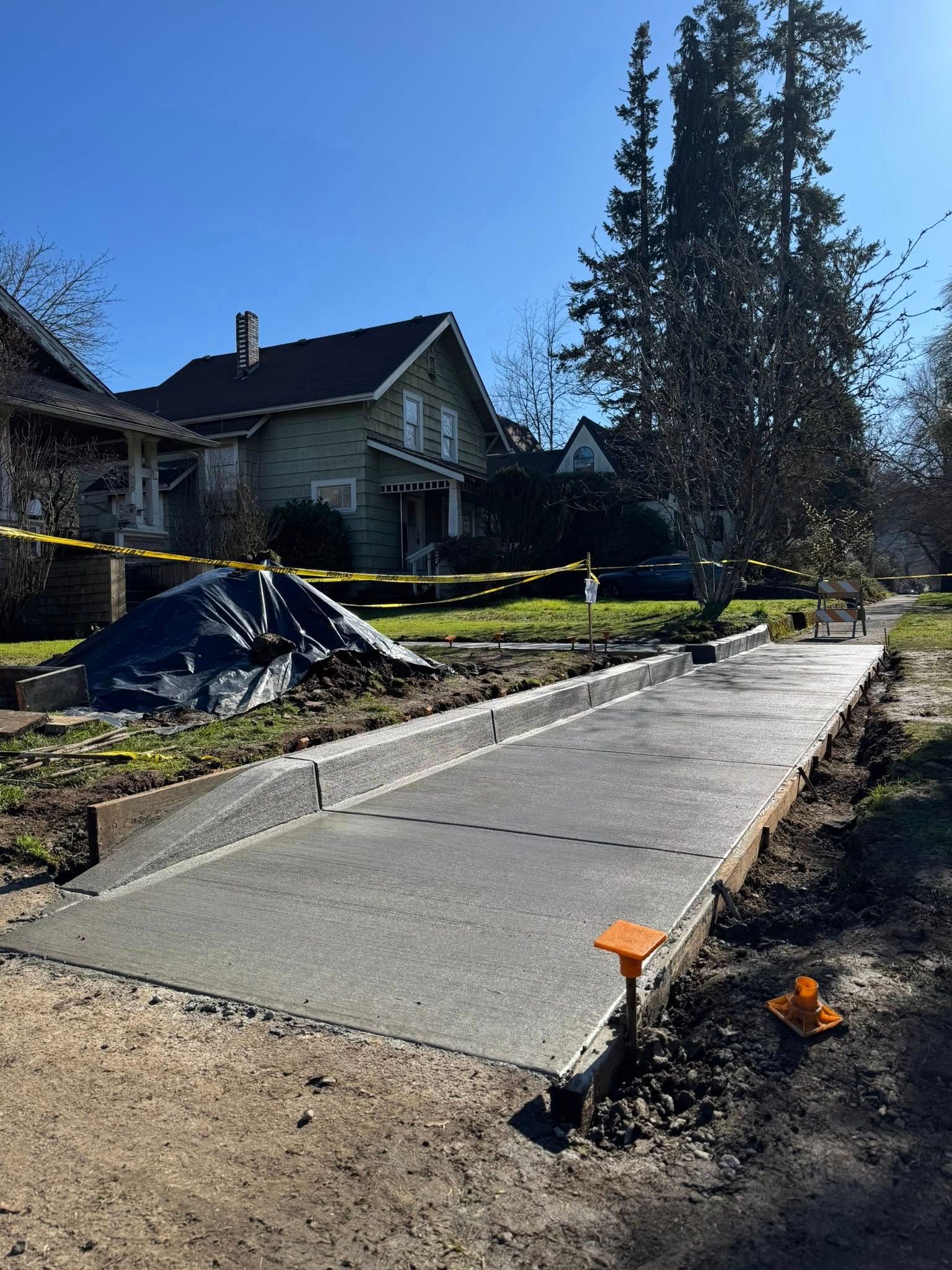 Newly poured concrete sidewalk and curb in a suburban neighborhood on a sunny day, marked off with yellow caution tape.