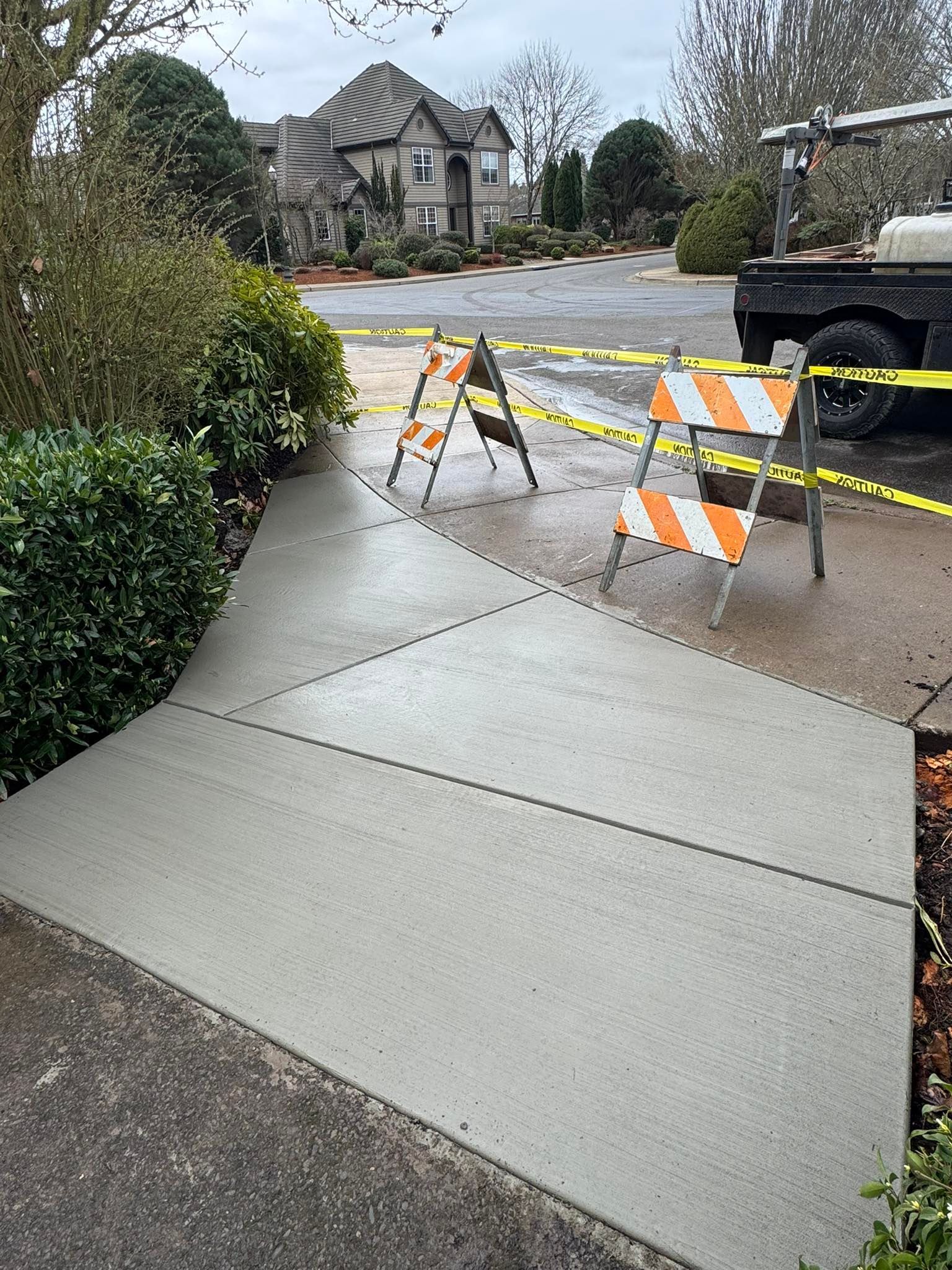 A newly poured concrete walkway blocked off by orange and white construction barricades and yellow caution tape.